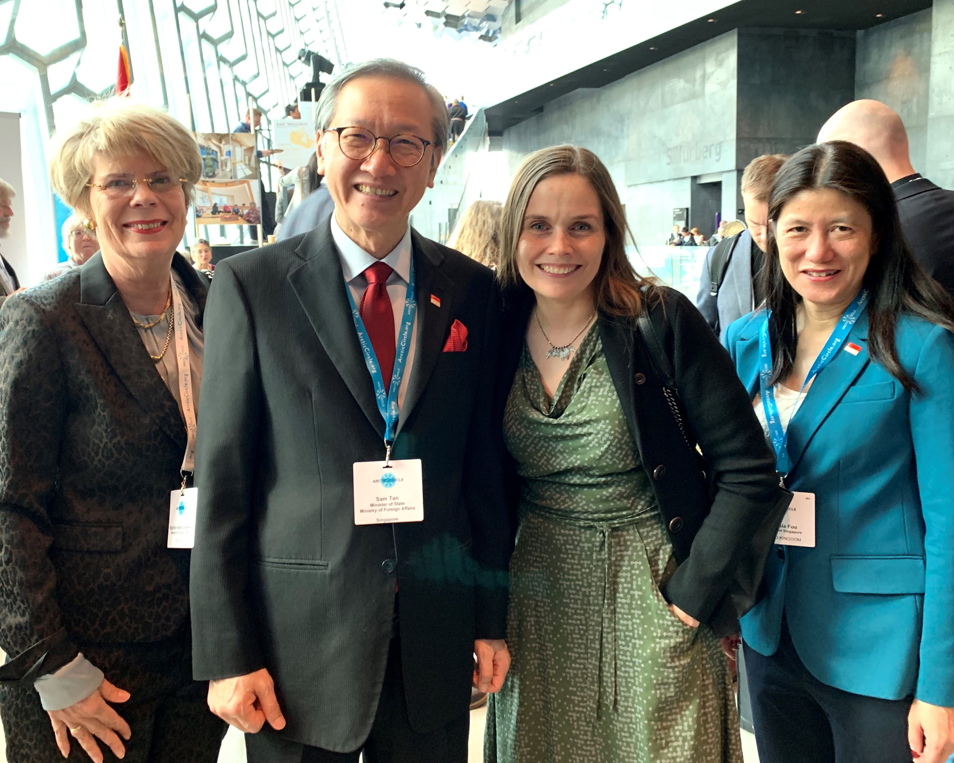 Four people pose indoors, wearing formal attire and lanyards. Sam Tan identified on a tag.